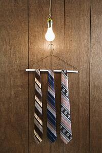 Three retro ties hanging on a wire hanger against wood paneling with a glowing lightbulb hanging down from above.