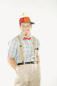 Young man dressed like nerd wearing propeller cap with hands in pockets looking at viewer with big smile