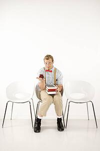 Young man dressed like nerd looking at viewer sitting with books and holding out apple