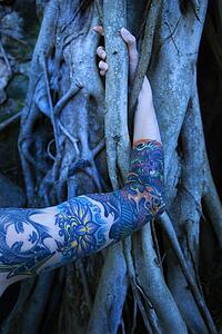 Blue-toned portrait of tattooed woman's arm intertwined with Banyan tree 