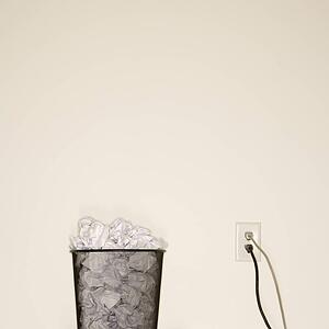 Full wire mesh trash can with crumpled paper next to electrical outlet and plugs