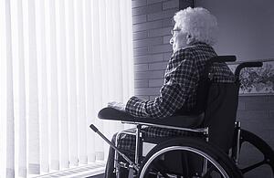Senior woman sitting in wheelchair looking out of window with blinds
