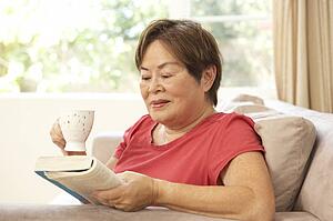 Senior Woman Reading Book With Drink At Home