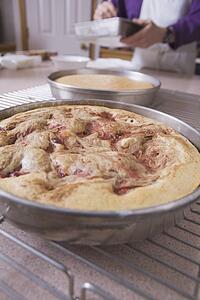 baked cake in a round pan on the cooling rack with baker in the background