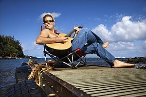 Young Man Sitting Lakeside and Playing Guitar