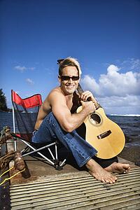 Young Man Sitting Lakeside and Holding Guitar