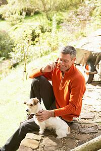 Man Outdoors On Mobile Phone With Dog Whilst On Break From Gardening