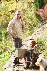 Man Outdoors On Mobile Phone With Dog Whilst On Break From Gardening