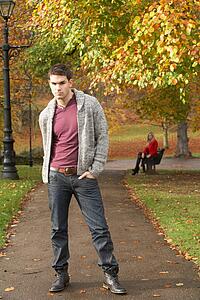 Teenage Boy Standing In Autumn Park With Female Figure On Bench In Background