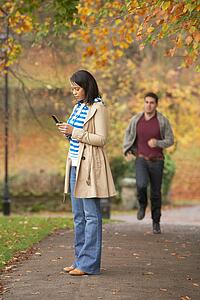 Teenage Girl Making Mobile Phone Call With Boyfriend Running Towards Her In Background