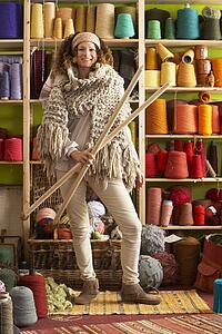 Woman Wearing Knitted Scarf Standing In Front Of Yarn Display Holding Giant Needles