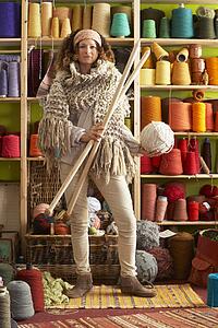 Woman Wearing Knitted Scarf Standing In Front Of Yarn Display Holding Giant Needles