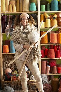 Woman Wearing Knitted Scarf Standing In Front Of Yarn Display Holding Giant Needles And Ball Of Wool