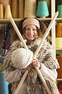 Woman Wearing Knitted Scarf Standing In Front Of Yarn Display Holding Giant Needles And Ball Of Wool