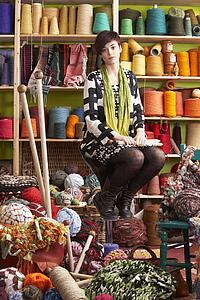 Young Woman Sitting On Stool Holding Knitting Needles In Front Of Yarn Display