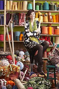 Young Woman Sitting On Stool Holding Knitting Needles In Front Of Yarn Display