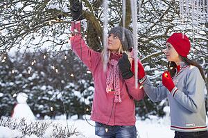Two Teenage Girls Hanging Fairy Lights In Tree With Icicles In Foreground