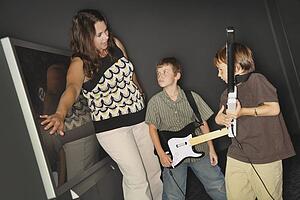 A Mother In Front Of The Television Screen Telling Her Boys To Stop Playing Their Music Game