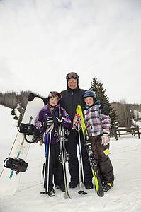 red deer, alberta, canada; a father and two daughters at a ski area holding a snowboard and skiis