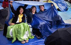 three ladies at an outdoor music concert (folkfest) protecting themselves under tarps from the rain; edmonton, alberta, canada