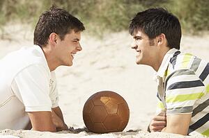 Two Young Men Relaxing On Beach With Football Together