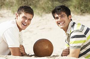 Two Young Men Relaxing On Beach With Football Together