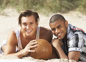 Two Young Men Relaxing On Beach With Football Together