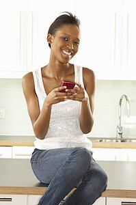 Woman Using Mobile Phone Sitting In Modern Kitchen 