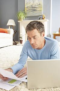 Man Using Laptop To Manage Household Bills Laying On Rug At Home 