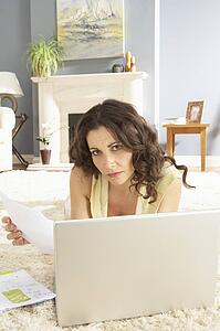 Woman Using Laptop To Manage Household Bills Laying On Rug At Home 