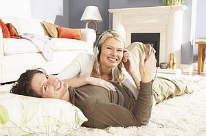 Couple Listening To MP3 Player On Headphones Relaxing Laying On Rug At Home 