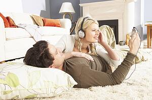Couple Listening To MP3 Player On Headphones Relaxing Laying On Rug At Home 