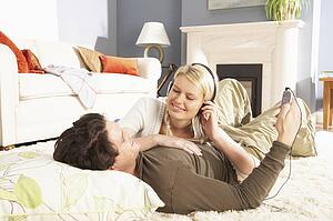 Couple Listening To MP3 Player On Headphones Relaxing Laying On Rug At Home 