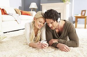Couple Listening To MP3 Player On Headphones Relaxing Laying On Rug At Home 