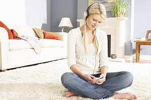 Woman Listening To MP3 Player On Headphones Relaxing Sitting On Rug At Home 