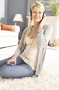 Woman Listening To MP3 Player On Headphones Relaxing Sitting On Rug At Home 