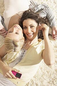 Two Women Listening To MP3 Player On Headphones Together Relaxing Laying On Rug At Home 