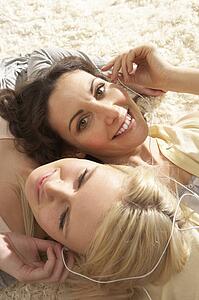 Two Women Listening To MP3 Player On Headphones Together Relaxing Laying On Rug At Home 