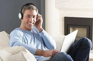 Man Using Laptop Wearing Headphones Relaxing Sitting On Sofa At Home 