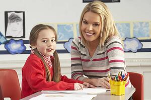 Female Primary School Pupil And Teacher Working At Desk In Classroom