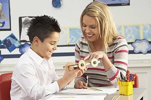 Male Primary School Pupil And Teacher Working At Desk In Classroom