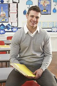 Portrait Of Male Teacher Sitting At Desk In Classroom