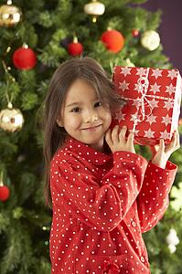 Young Girl Holding Wrapped Present In Front Of Christmas Tree