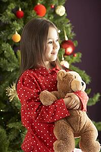 Young Girl Cuddling Teddy Bear In Front Of Christmas Tree