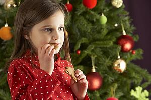 Young Girl Eating Star Shaped Christmas Cookie In Front Of Christmas Tree