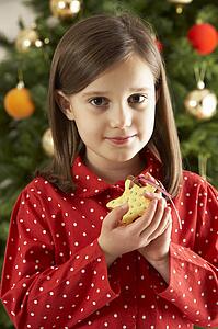 Young Girl Eating Star Shaped Christmas Cookie In Front Of Christmas Tree