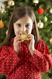 Young Girl Eating Star Shaped Christmas Cookie In Front Of Christmas Tree