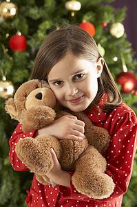 Young Girl Cuddling Teddy Bear In Front Of Christmas Tree