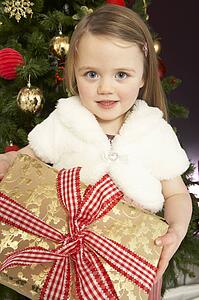 Young Girl Holding Gift In Front Of Christmas Tree