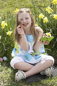 Girl Eating Chocolate Egg On Easter Egg Hunt In Daffodil Field